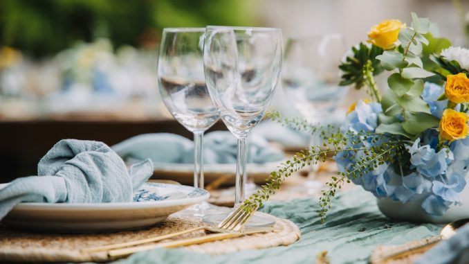 Elegancko nakryty stół na świeżym powietrzu; na pierwszym planie stoją dwa kieliszki do wina, obok talerze z błękitnymi, materiałowymi serwetkami. Dekorację uzupełnia bukiet z żółtych róż i niebieskich hortensji, tworząc świeży, wiosenno–letni klimat.